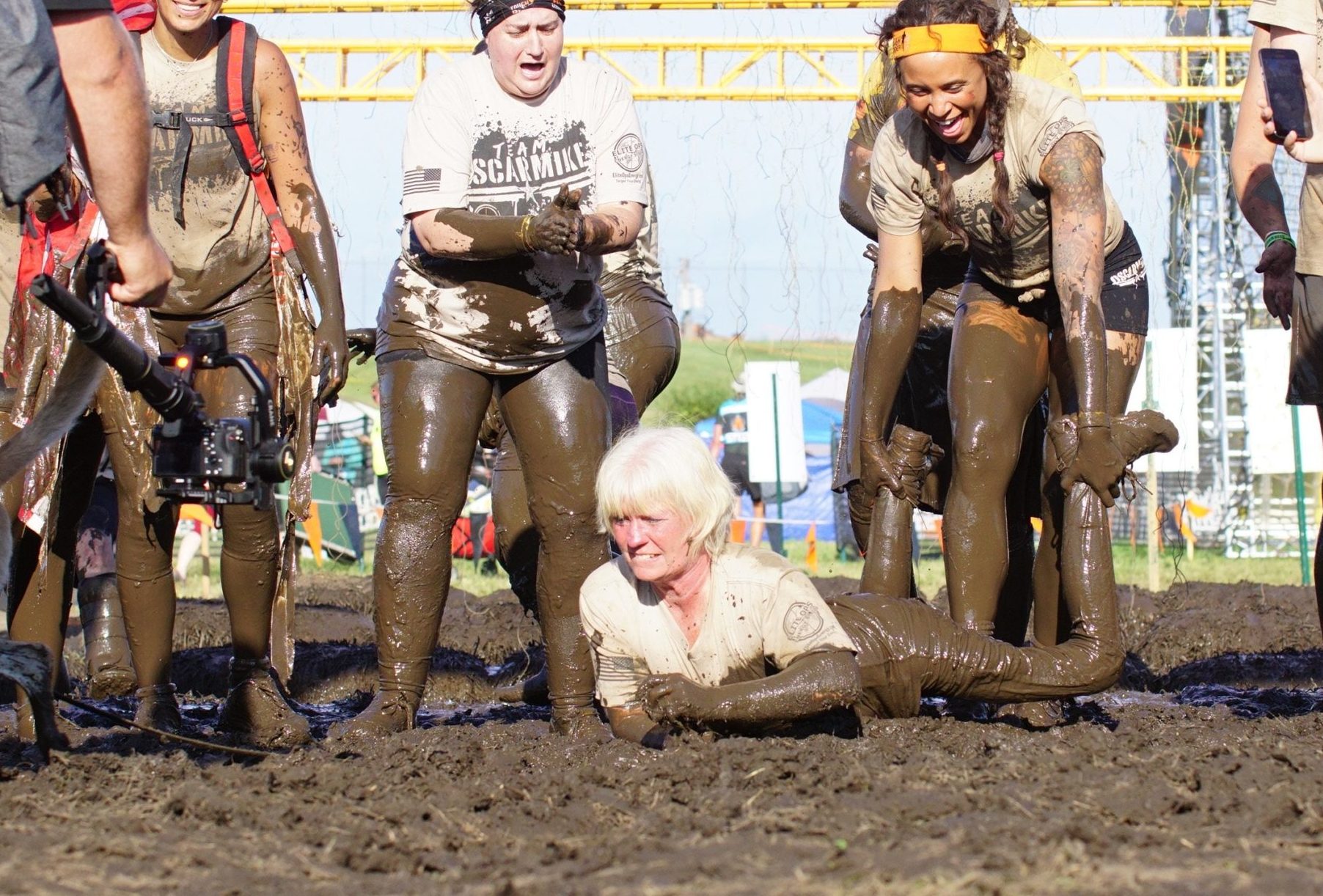Old lady participant crawling through the mud while here teammates cheering for her